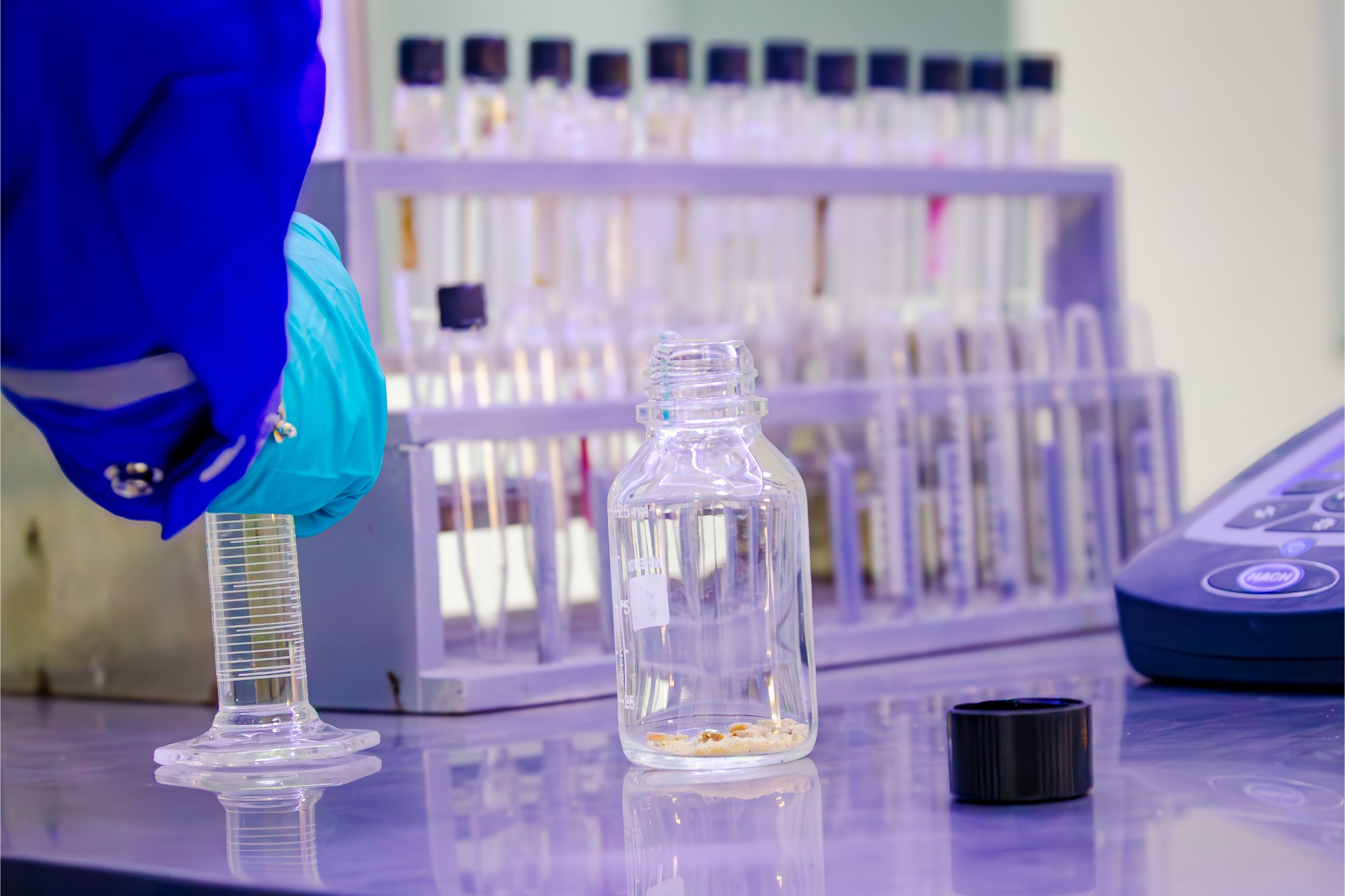 Gloved hand holding a graduated cylinder beside a sample bottle
